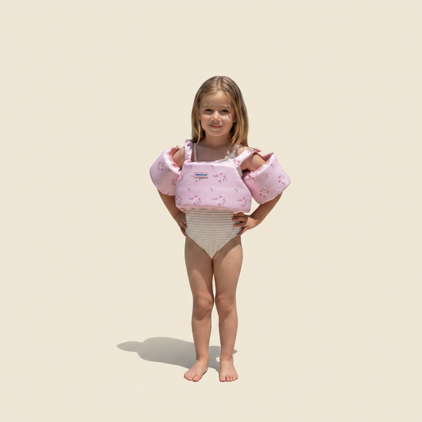 Young girl holding pink swim floaties by a pool with greenery in the background