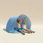 Two children sitting inside a blue beach tent with cartoon designs on a beige background